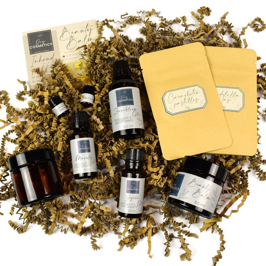 Alle ingrediënten, cacaoboter, candelillawas, jojoba olie, zonnebloemolie, marula olie, vitamine E en geranium etherische olie, van het DIY beautybalsem pakket bij elkaar op papierkrullen.